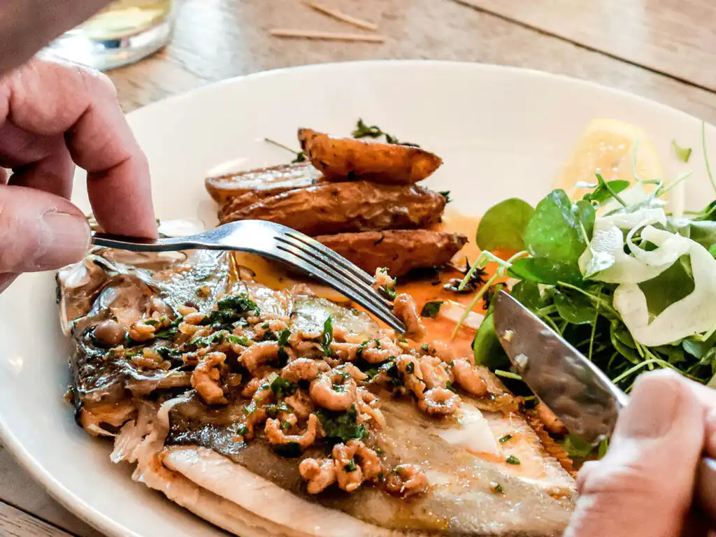 A person cuts a piece of fish with a fork, enjoying a seafood dish at Mawgan Porth Restaurant.