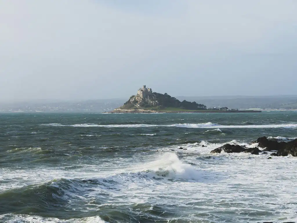 Stormy ocean near a rocky shore, with St Michael's Mount visible in the background, near Marazion, a great place for food and drink in Cornwall.