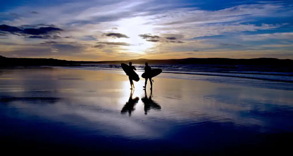Two surfers stand on a Cornish beach at dusk, holding their surfboards, ready to catch the evening waves as part of a lesson at a West Cornwall Surf School.