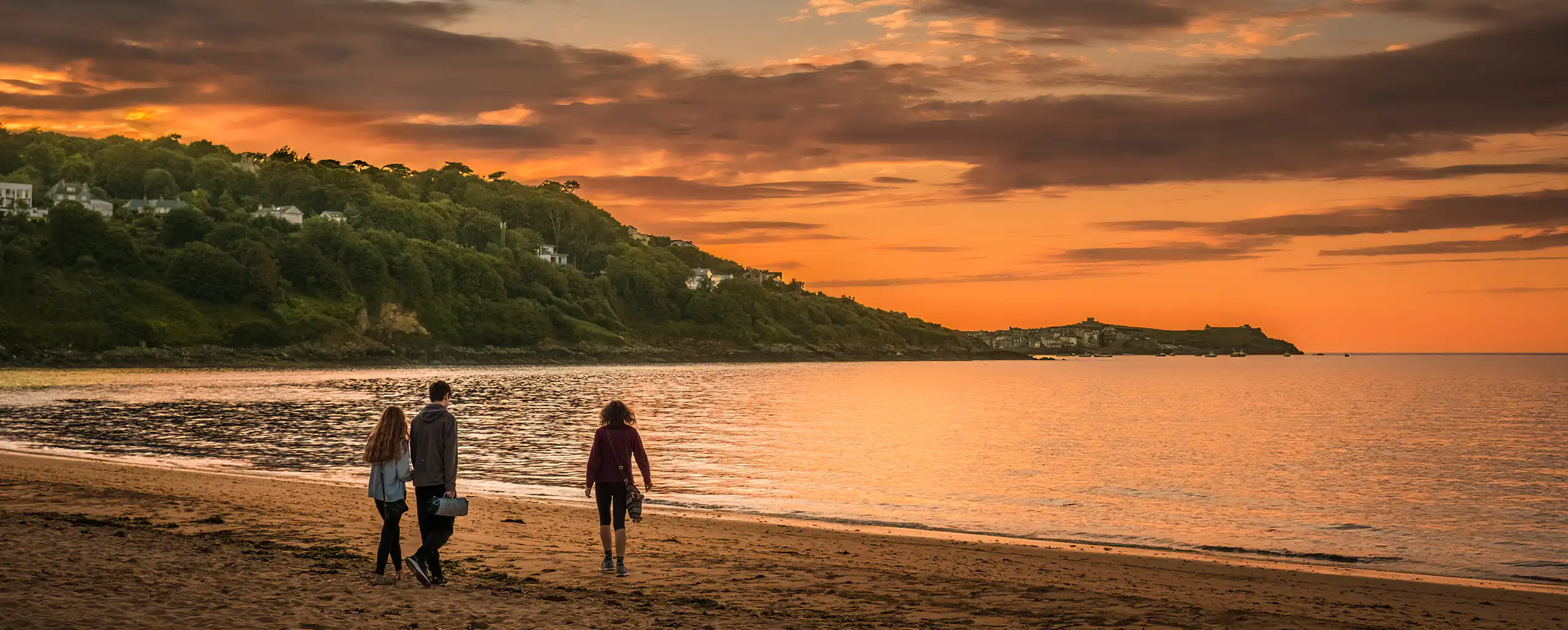 Three people stroll along Carbis Bay Beach at sunset, with vibrant colors reflecting on the water and soft sand beneath their feet, a great place for food in Carbis Bay.