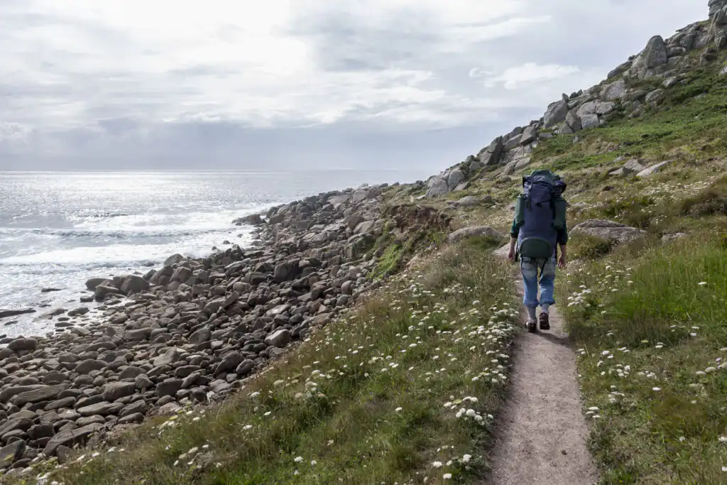A woman with a backpack walks along the South West Coastal Path, with the ocean visible beside her in Cornwall, part of the Salt Path route.