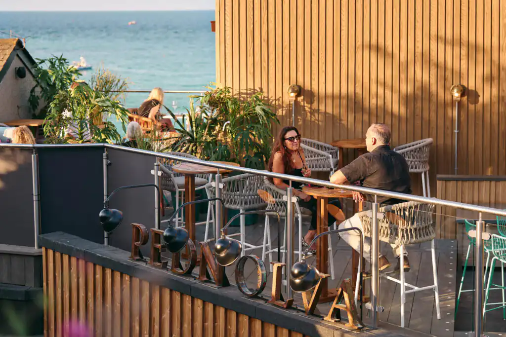 A couple enjoys a meal on a restaurant balcony, overlooking the ocean at Pedn Olva in St Ives, Cornwall, a great pub with a beer garden in Cornwall.