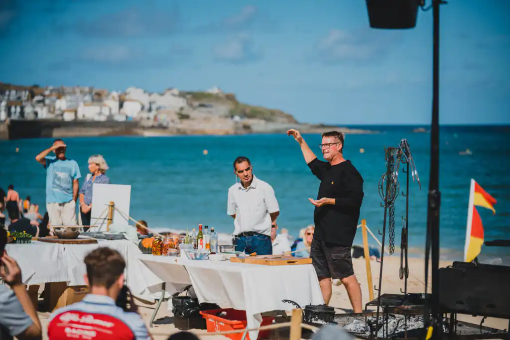 A man demonstrates cooking at St Ives Food Festival, with a table on the beach and an audience watching nearby, a great Cornish summer festival.