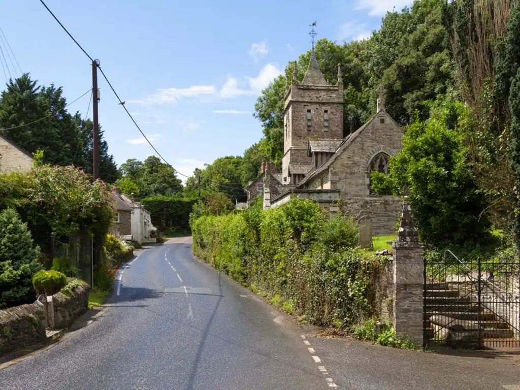 A scenic country road lined with trees, featuring a quaint church nestled beside it in Little Petherick village, a great piece of arts and heritage in Padstow.