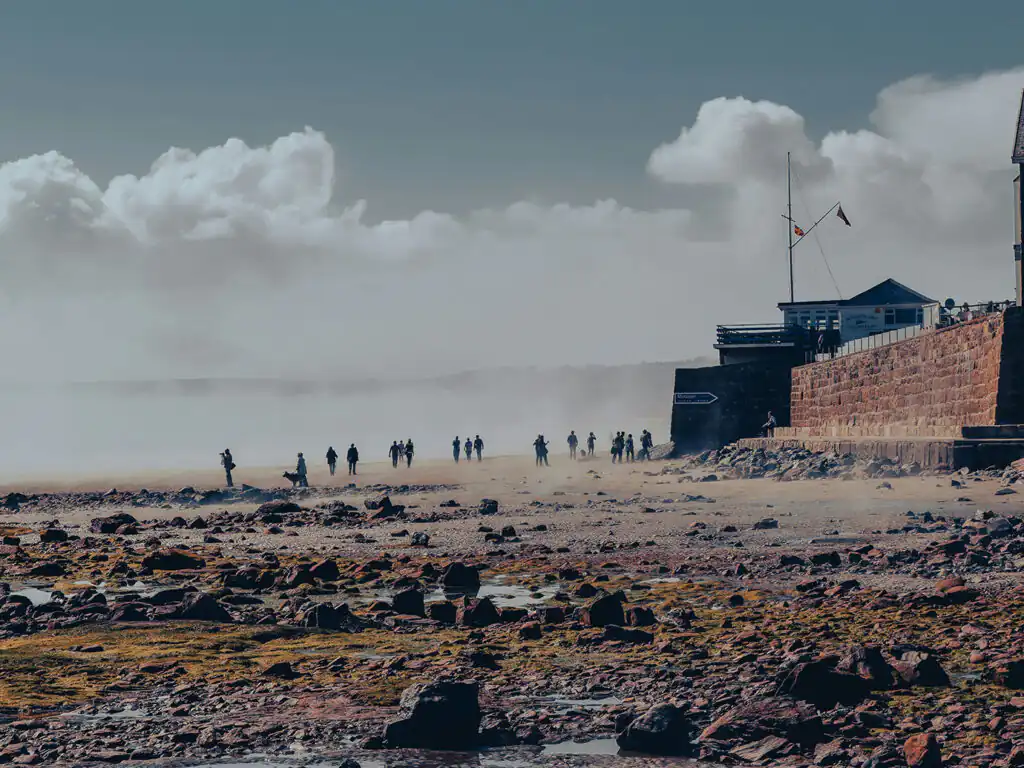 A cloudy sky looms over Marazion Beach, where people enjoy the seaside atmosphere and activities, adjacent to some great food and drink locations in Marazion.