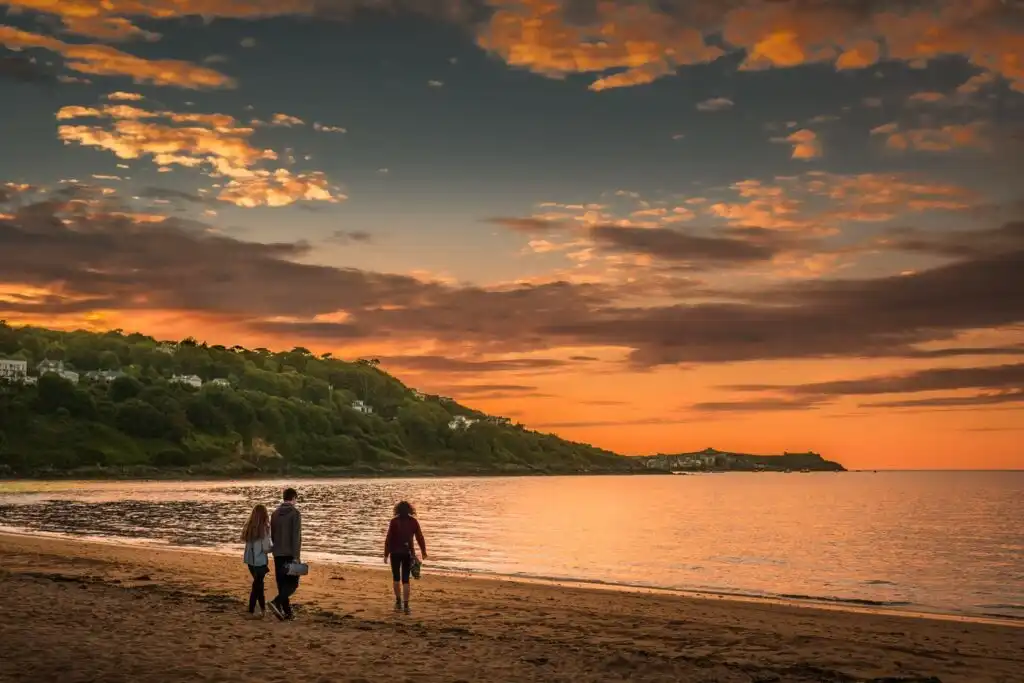 Three people stroll along Carbis Bay Beach at sunset, with golden hues reflecting on the calm water, a must visit when planning a holiday in Carbis Bay.