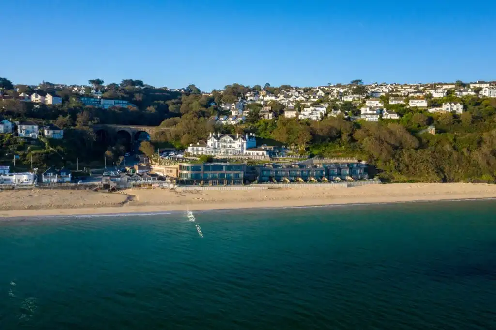 Aerial view of Carbis Bay Hotel, showcasing the beach and hillside houses under a clear blue sky, a great place for food and drink in Carbis Bay.
