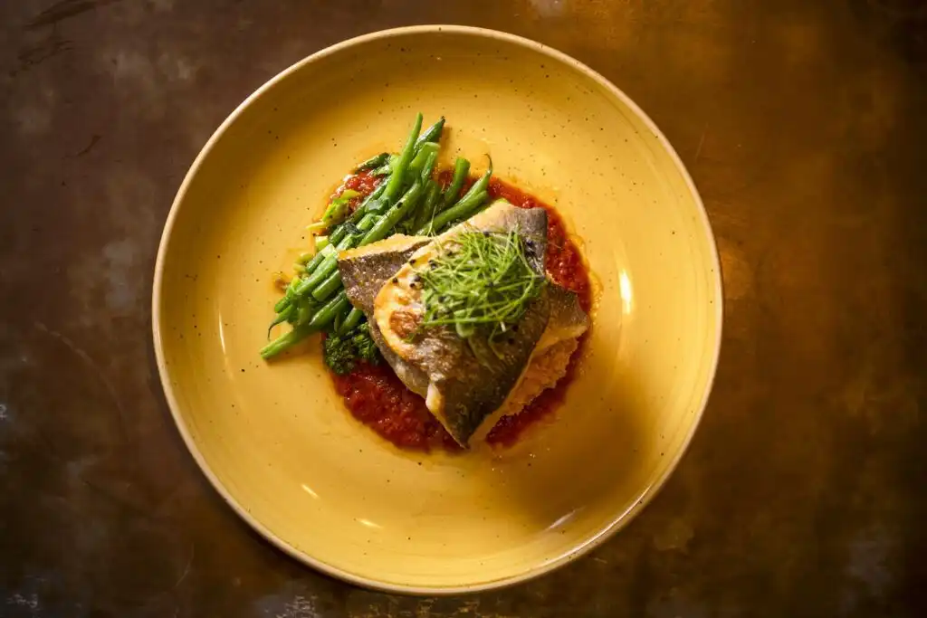 A colourful fish dish served on a yellow plate, garnished with green beans and tomatoes, from The Gannet in Carbis Bay, a great place for food and drink.